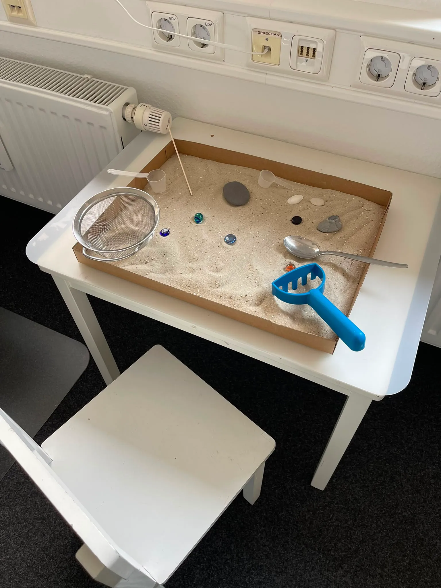 Kleiner Tisch mit Sandkasten, Sieb, Löffel und Steinen